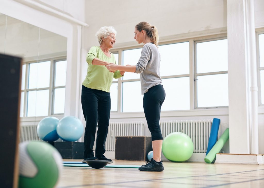 Female trainer helping senior woman in a gym exercising with a bosu balance training platform. Elder woman being assisted by gym instructor while workout session.