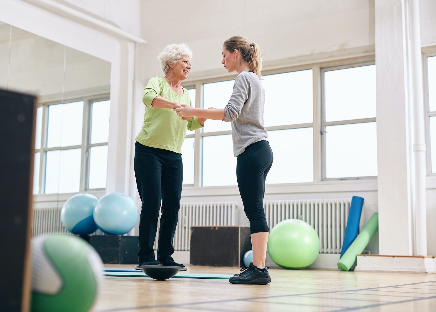 Female trainer helping senior woman in a gym exercising with a bosu balance training platform. Elder woman being assisted by gym instructor while workout session.
